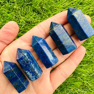May include: Six blue lapis lazuli crystal points, also known as lapis lazuli towers, are arranged in a hand. The points are various sizes and have a dark blue colour with gold flecks.