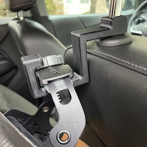 May include: A black plastic car seat headrest hook with a gray buckle and a black strap. The hook is attached to the headrest of a car seat.