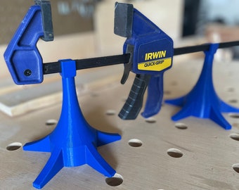 May include: Blue Irwin Quick-Grip clamps with black bars and yellow text, supported by blue 3D-printed stands. The clamps are on a light brown wooden surface with evenly spaced holes, suggesting a workshop setting.