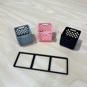 May include: Three small plastic crates, one gray, one pink, and one black, are arranged on a wooden surface. A black plastic frame with three square openings is below the crates.