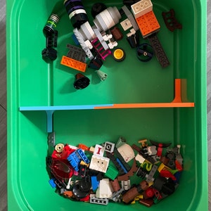 May include: A green plastic bin filled with assorted colorful toy building blocks. The blocks are various shapes and sizes, including orange, white, black, and blue pieces. Some blocks are assembled into larger structures, while others are loose. A blue and orange track is also visible.