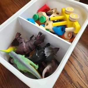 May include: A white plastic bin divided into two sections, filled with toys. One side contains colorful wooden blocks and a yellow toy car. The other side holds various plastic dinosaur figures in shades of green, brown, and black.