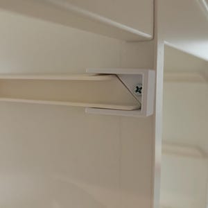 May include: White plastic shelf bracket with a screw visible. The bracket is attached to a white shelf.