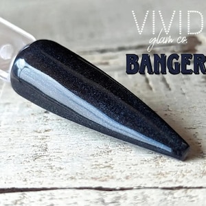 Könnte beinhalten: Ein schwarzer Nagellack-Swatch mit dem Wort "BANGER" in dunkelblauen Blockbuchstaben. Der Nagellack befindet sich auf einem durchsichtigen Plastikstab, und der Hintergrund ist eine hellfarbige Holzoberfläche. Die Worte "VIVID glam co." stehen über dem Wort "BANGER".