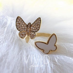 May include: Gold ring featuring two butterfly designs. One butterfly is set with clear rhinestones, while the other is white enamel.