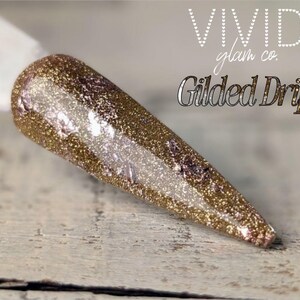 May include: A gold glitter nail with a pointed tip, displaying a shimmering effect. The nail is on a white stick, with the words "VIVID glam co." and "Gilded Drip" in the background.