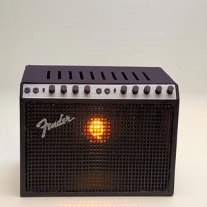 May include: A black Fender amplifier with a grid-like speaker cover and silver knobs. The word "Fender" is displayed on the front. The amplifier has a glowing orange light in the center, suggesting it is turned on.