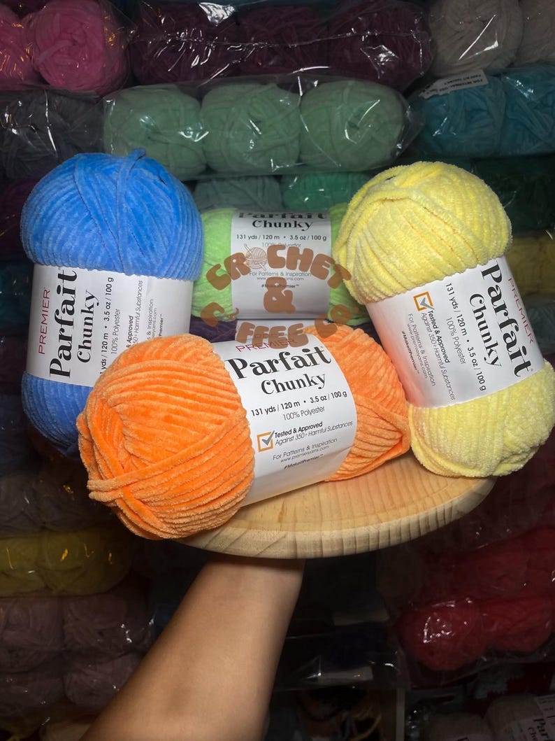 Puede incluir: Surtido de ovillos de lana de colores en varios tonos de azul, naranja, amarillo y verde. La lana est&aacute; etiquetada como "Parfait Chunky" y es 100% poli&eacute;ster. La lana tiene 131 yardas por 120 metros y 3,5 onzas por 100 gramos.