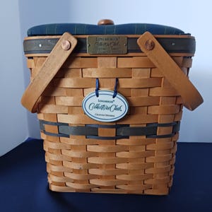 May include: Woven brown basket with wooden handles and a blue and green plaid padded lid. Features a metal plaque and a hanging oval tag that reads "Collectors Club". The basket has a dark gray band around the middle.