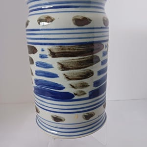 May include: A white ceramic vase with blue and brown stripes and abstract designs. The vase has a rounded base and a cylindrical body.