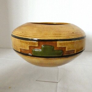 May include: A ceramic bowl with a tan exterior and a dark brown and green geometric design. The bowl has a rounded shape and a wide opening. The design includes a series of rectangular and stepped shapes. The bowl is displayed on a clear stand.