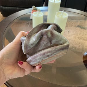 May include: A handcrafted, ceramic, abstract-shaped bowl with a textured surface. The bowl is primarily a muted purple color with hints of green and white. The bowl is held in a hand, with candles in the background.