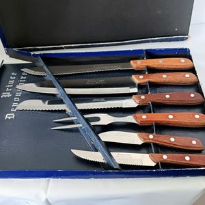 May include: A set of eight stainless steel kitchen knives with wooden handles in a blue box. The knives include a bread knife, a carving knife, a chef's knife, a paring knife, a utility knife, and a steak knife. The box has the words "Kitchen Pride" printed on it.