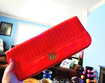 May include: A bright red clutch with a glossy finish. The rectangular bag features a gold-colored emblem and vertical stitched detailing. The clutch is held at an angle, showcasing its design and color.