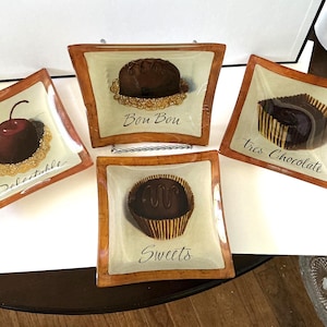 May include: A set of four square glass plates with brown borders, each featuring a different chocolate dessert illustration and text. The plates display a chocolate-covered cherry labeled Delectable, a truffle labeled Bon Bon, a chocolate square labeled tres Chocolate, and a truffle labeled Sweets.