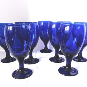 May include: A collection of eight cobalt blue wine glasses. Each glass features a rounded bowl, a slender stem, and a circular base. The glasses are arranged on a white surface, showcasing their reflective qualities and deep color.