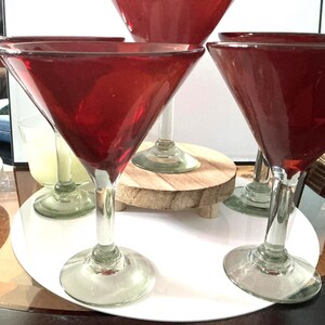 May include: A set of four martini glasses with deep red, conical bowls and clear glass stems and circular bases. The glasses are arranged in a cluster, with one glass positioned slightly higher than the others on a small wooden riser.