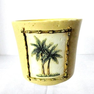 May include: A light yellow ceramic planter with a rectangular inset featuring a relief of two palm trees. The inset is framed by a bamboo-style border. The planter has a slightly flared shape.