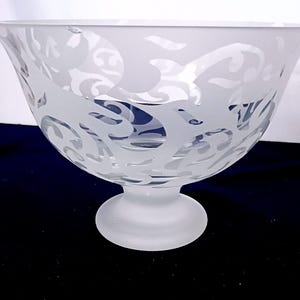 May include: A frosted glass bowl with a wide, flared rim and a pedestal base. The bowl features a decorative, etched pattern of swirling shapes. The bowl is made of clear glass with a frosted finish, creating a soft, diffused appearance.