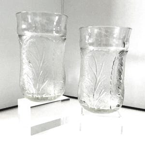 May include: Two clear glass tumblers featuring a textured surface with a repeating embossed leaf pattern. Each glass has a slightly flared rim and a rounded base, showcasing a translucent design that catches the light across the glass body.