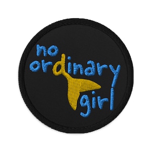Può includere: Un cerotto rotondo nero con ricamo blu e giallo. Il testo "no ordinary girl" è ricamato in blu con una coda di pesce stilizzata gialla.