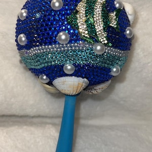Può includere: Un sonaglio decorativo con una sfera blu tempestata di gioielli, ornata di perle, conchiglie e un pesce di strass. L'impugnatura è azzurra. Un oggetto unico e fatto a mano.