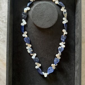 May include: A necklace made of blue and white stone beads in the shape of leaves. The necklace is displayed on a black velvet background.