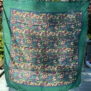 May include: A green silk scarf with a floral pattern of roses and smaller flowers in shades of yellow, blue, and purple. The scarf has a fringed edge.