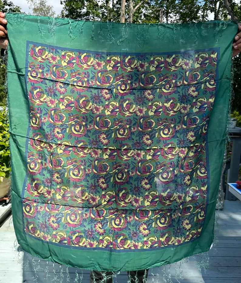 May include: A green silk scarf with a floral pattern of roses and smaller flowers in shades of yellow, blue, and purple. The scarf has a fringed edge.