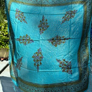 May include: A turquoise blue silk scarf with a geometric pattern and a gold border. The scarf has a fringe on the edges.
