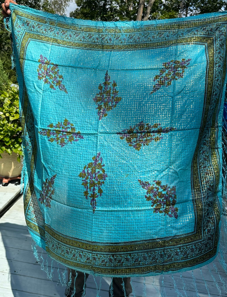 May include: A turquoise blue silk scarf with a geometric pattern and a gold border. The scarf has a fringe on the edges.