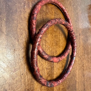 May include: Two red leather rings with a snakeskin pattern. The rings are intertwined and are on a wooden surface.
