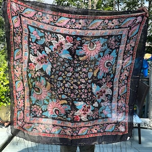 May include: A black silk scarf with a floral pattern in shades of pink, blue, and gold. The scarf has a fringed edge and is made of a lightweight fabric.