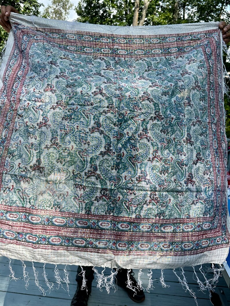 May include: A white cotton throw blanket with a paisley pattern in shades of blue, green, and red. The blanket has a fringe border and is woven with a grid pattern.