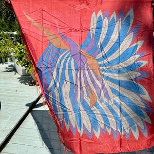May include: A red silk scarf with a colorful bird design. The bird has blue, white, purple, and brown feathers. The scarf has fringe on the edges.
