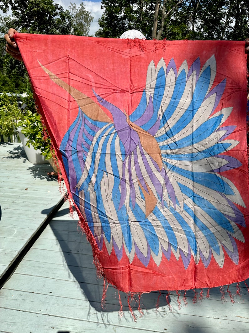 May include: A red silk scarf with a colorful bird design. The bird has blue, white, purple, and brown feathers. The scarf has fringe on the edges.