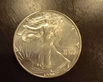 Könnte beinhalten: Eine silberne American Eagle-Münze mit dem Jahr 1990 auf der Unterseite. Die Münze zeigt eine Darstellung von Lady Liberty, die mit einer Fackel und einem Olivenzweig geht.