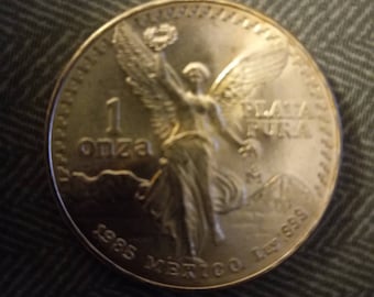 May include: A silver coin featuring a winged figure holding a laurel wreath. The coin reads "1 onza" and "Plata Pura" with the year "1985" and "Mexico Ley .999" inscribed. The coin is round and has a detailed design.