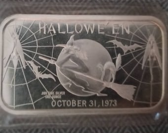 May include: A silver bar with a Halloween theme. It features a witch flying on a broomstick in front of a full moon with a spider web background. The text "HALLOWEEN" is at the top and "OCTOBER 31, 1973" is at the bottom. The bar also says "999 FINE SILVER ONE OUNCE".