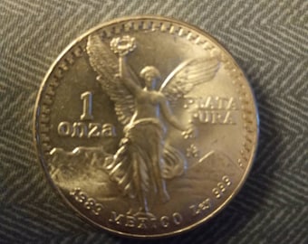 May include: A silver coin with an image of a winged figure holding a torch. The coin reads "1 onza" and "Plata Pura" with the year "1983" and "Mexico Ley .999" inscribed around the edge. The coin is on a textured gray surface.