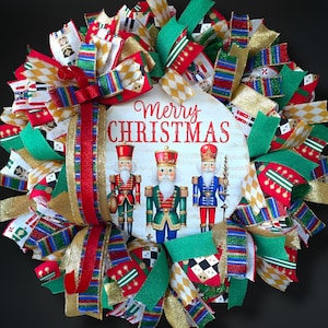 May include: A festive Christmas wreath with a wooden sign that reads "Merry Christmas" and features three nutcracker figures. The wreath is adorned with red, green, gold, and white ribbons, creating a colorful and cheerful holiday decoration.