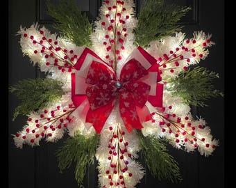 Op de afbeelding: Een sneeuwvlokvormige krans met witte klatergoudtakken, groene dennentakjes en rode bessentakken. Het midden is versierd met een grote rood-witte strik met sneeuwvlokpatroon en het geheel is verlicht met kleine lampjes.