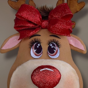 May include: A festive reindeer head wreath with brown antlers, a red glitter bow, and a red glitter nose. The reindeer has large eyes with long eyelashes and a smiling mouth. The wreath is a holiday decoration.