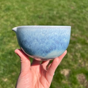 May include: A handmade ceramic bowl with a spout, held in a hand. The bowl has a gradient of blue hues, transitioning from a lighter shade at the rim to a darker, mottled blue at the base. The bowl is likely for serving or mixing.