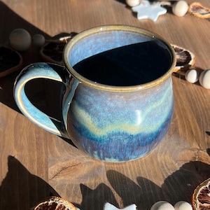 May include: A handmade ceramic mug with a blue and teal glaze. The mug has a rounded shape with a large handle and a light brown rim. The mug is approximately 4 inches tall and 4 inches in diameter. The mug is on a wooden surface.