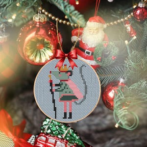 May include: A cross-stitch Christmas ornament featuring a mouse dressed as a king, holding a staff. The ornament is round, with a light blue background and a red bow. The ornament hangs on a Christmas tree with red ornaments and a Santa Claus figurine.