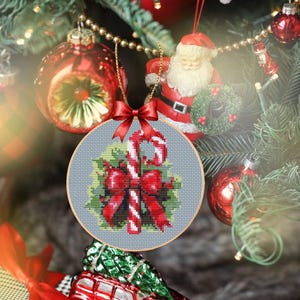 May include: A round, cross-stitched Christmas ornament with a candy cane and red bow design. The ornament is hanging on a Christmas tree with red and gold ornaments and a Santa Claus figurine. The ornament is approximately 3 inches in diameter.