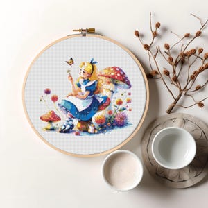 Può includere: Un telaio da ricamo a punto croce con una colorata illustrazione di Alice nel Paese delle Meraviglie. Alice, in un vestito blu, è seduta su un fungo, con una farfalla. Il disegno comprende altri funghi e fiori, il tutto all'interno di un telaio di legno.