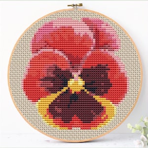 May include: A cross-stitch embroidery of a pansy flower in a wooden hoop. The flower features shades of red, pink, yellow, and brown, with a detailed, pixelated design. The embroidery is set against a neutral-colored fabric.
