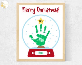 May include: A framed print featuring a snow globe illustration with a green handprint inside. The text Merry Christmas is at the top in red. A yellow star with 2026 is above the handprint, and a red base with the name Noah is at the bottom.